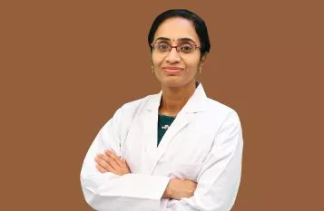 Female doctor with arms crossed, wearing a white lab coat and glasses, against a brown background.