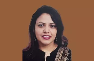 Portrait of a smiling woman with dark hair, wearing a black top and patterned scarf, against a brown background.