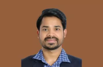 Portrait of a man with short dark hair and a beard, wearing a blue checkered shirt and dark blazer, facing the camera against a brown background.