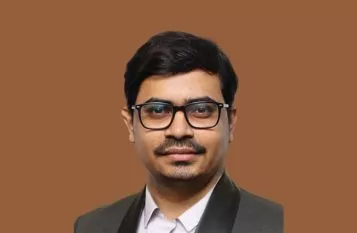 Portrait of a man in a dark suit and glasses, against a solid brown background.