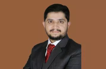 Professional head-and-shoulders portrait of a man in a dark suit and red tie against a brown background.